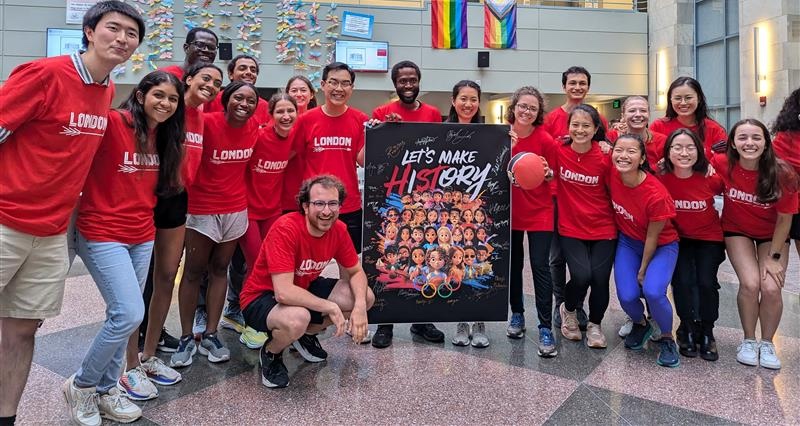 London Society students posing in the TMEC Atrium with their Society Olympics poster. The poster says "Let's Make HISTORY" with the letters H, S, and T standing out in a different color.