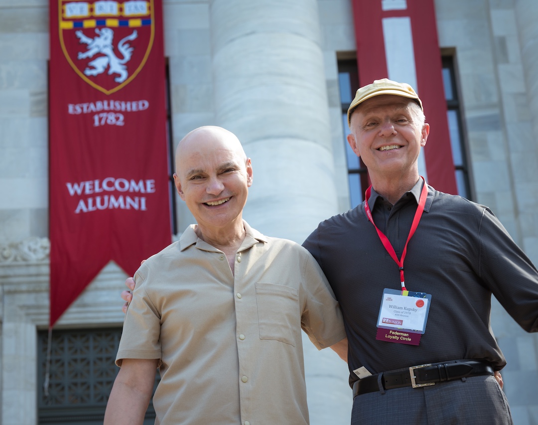 Two alumni pose in front of Gordon Hall with a "Welcome Alumni" banner hanging in the background.