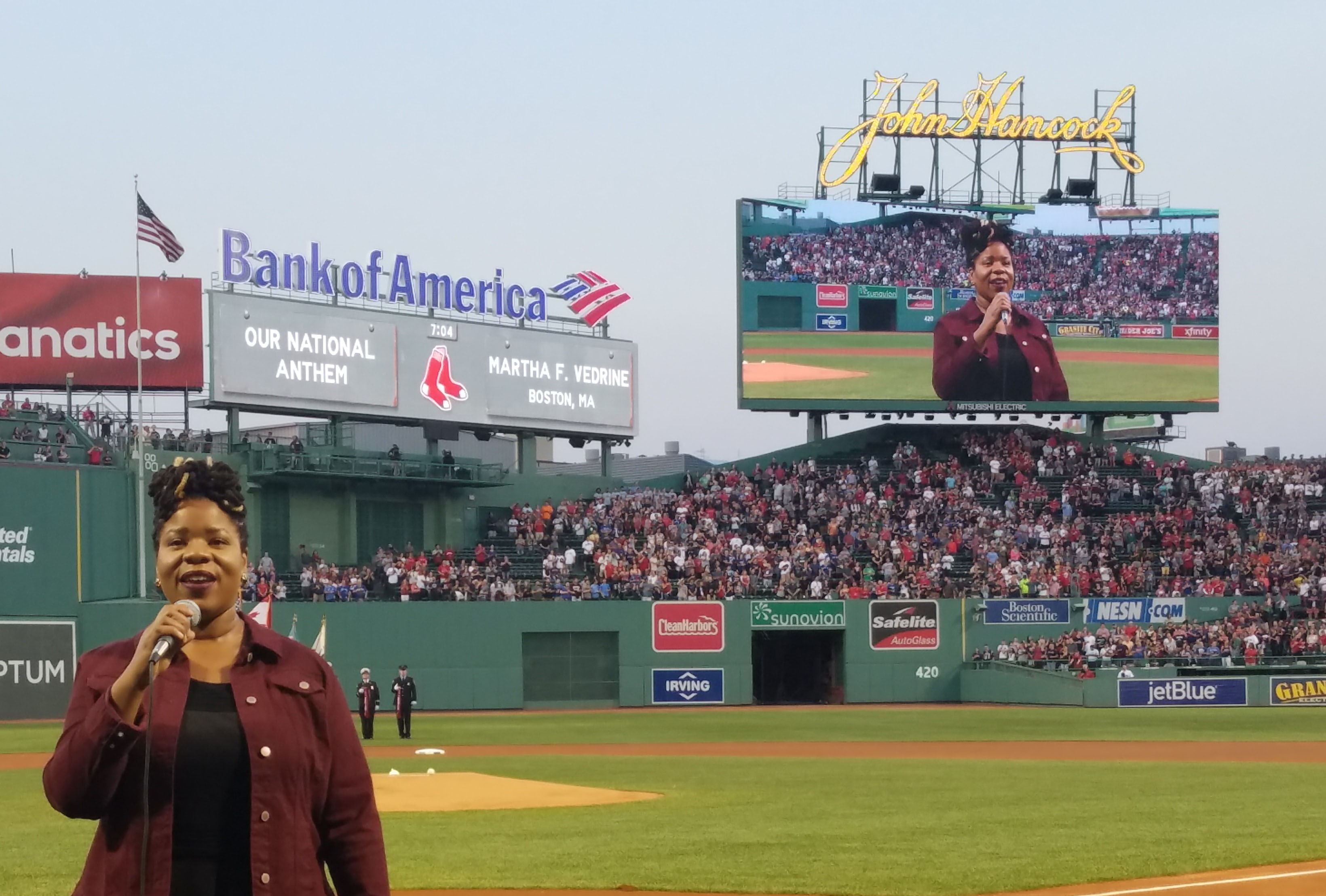 Martha Vedrine singing on the field at Fenway Park, with her image displayed on the jumbotron in the background