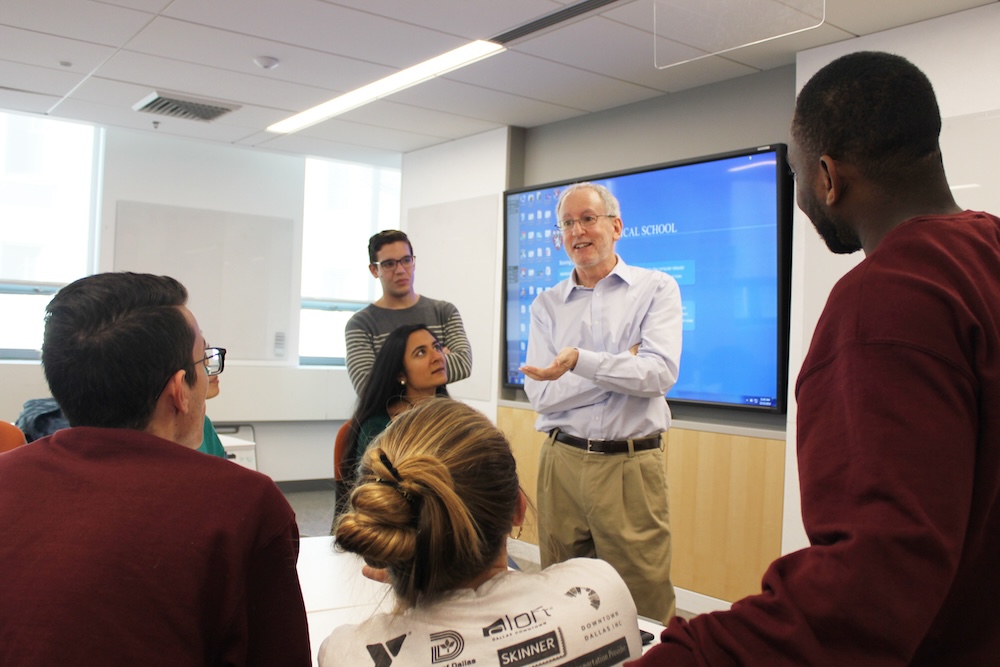 A small group discussion takes place around a table in a learning suite where a faculty member is with five students.