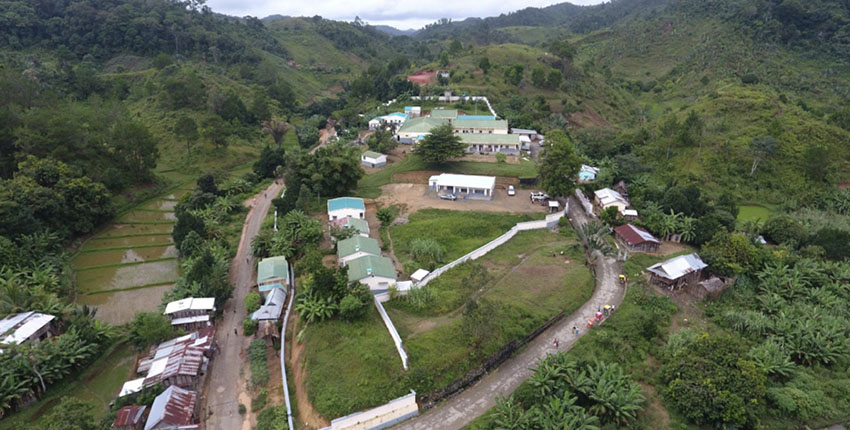 An aerial view of a hospital on an unpaved road surrounded by forested hills, farm fields, and small buildings. 