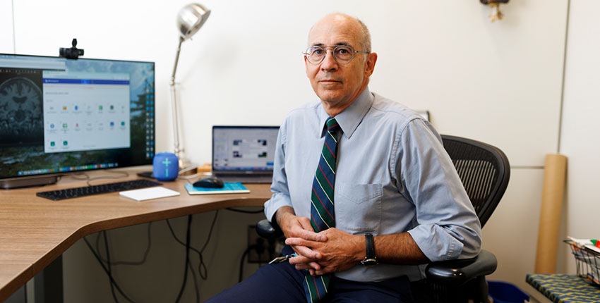 A man with clasped hands sits turned toward the camera at a desk. The computer monitor shows a brain scan.