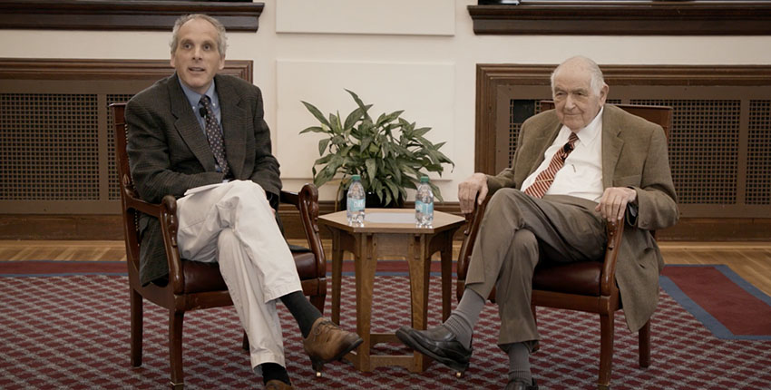 Two men in sports coats and ties sit in armchairs in front of a small table.