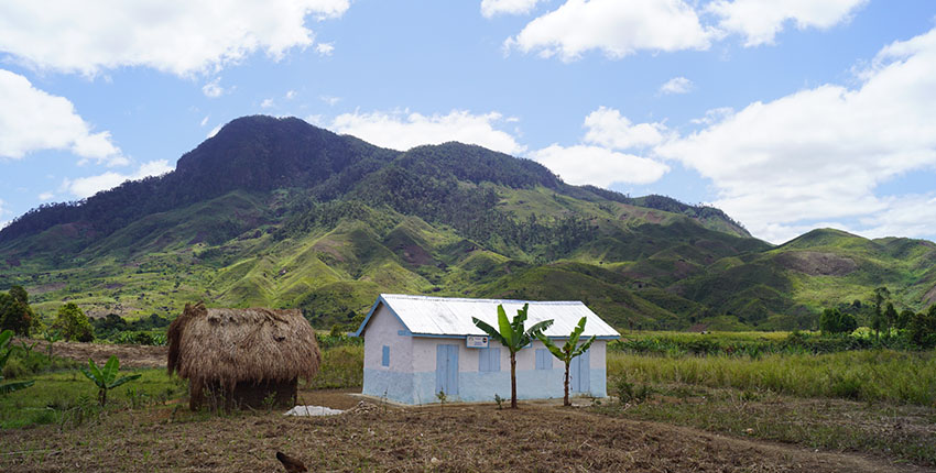 A small rural health center with white and light blue walls stands in front of a thatched hut in a wide, green valley surrounded by lush mountains.