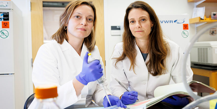 Two women in gloves and white lab coats stand at a lab bench as if caught mid-experiment, gazing at the camera with slight smiles