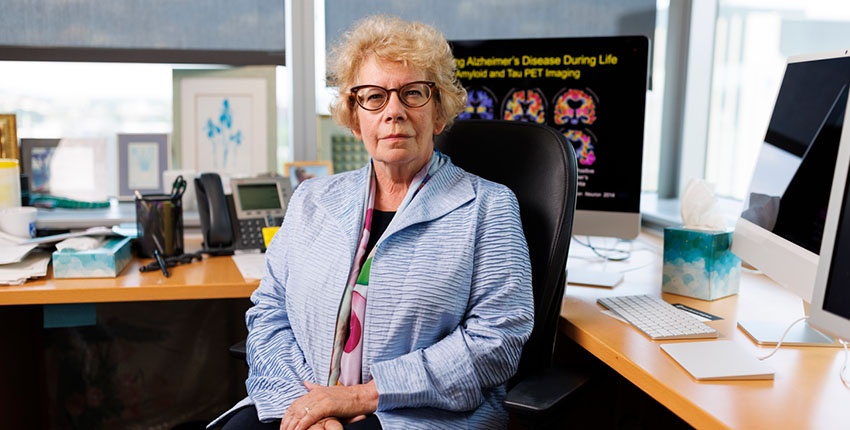 A woman sits with hands folded in front of a desk on which a computer shows several colorful brain images
