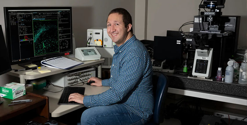 A man at an equipment-filled desk turns to smile at the camera