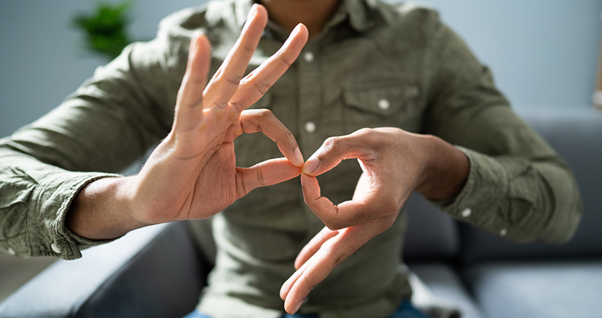 Close-up of two hands belonging to a person of color as they form the ASL sign for “interpreter.” The person’s head is unseen as they sit in a chair in a button-down shirt.