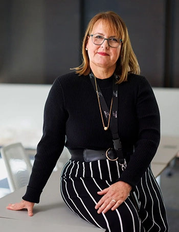 A woman in a dark top and striped pants smiles slightly at the camera while perched on a desk