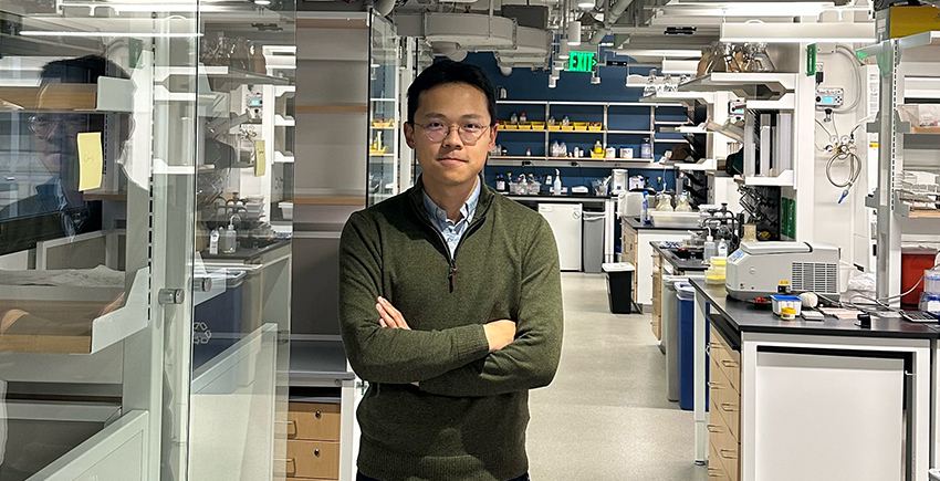 A man stands in a chemistry lab, with machinery on lab benches and reflections on glass partitions.