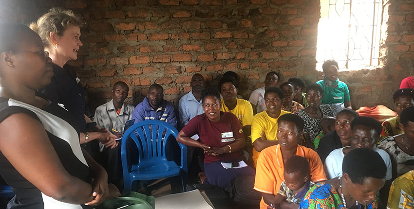 A group of adults seated closely together inside a simple brick-walled room listen attentively as two women stand at the front speaking. The audience includes men and women of different ages, some holding notebooks or bags, creating the feel of a community meeting or training session.