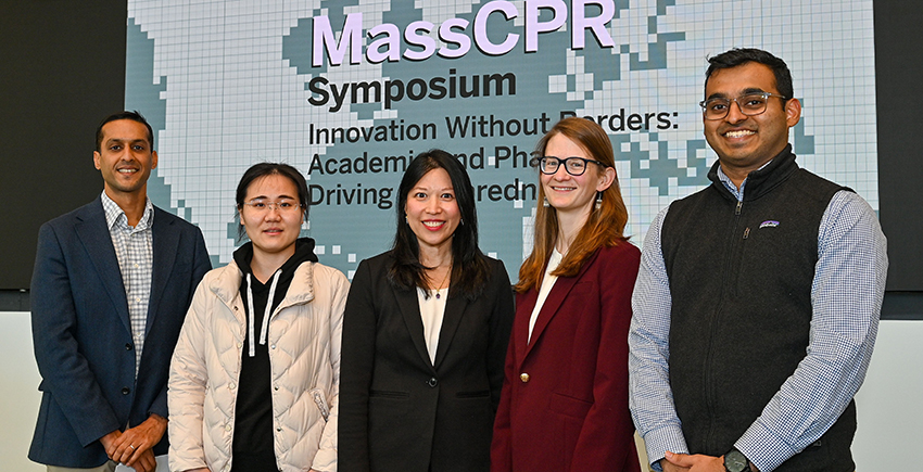 Five people in business clothes pose for a photo in front of a screen with the MassCPR symposium name