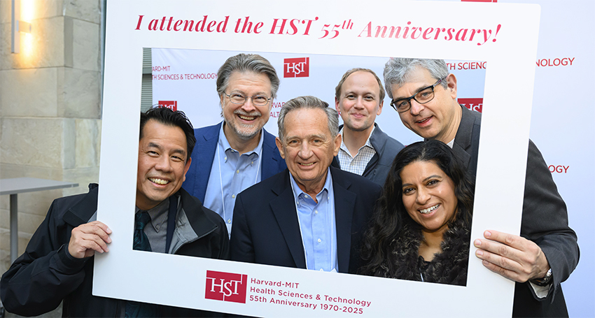 Six people pose, smiling, inside a large portable photo frame that says “Harvard-MIT Health Sciences & Technology 55th Anniversary 1970-2025”