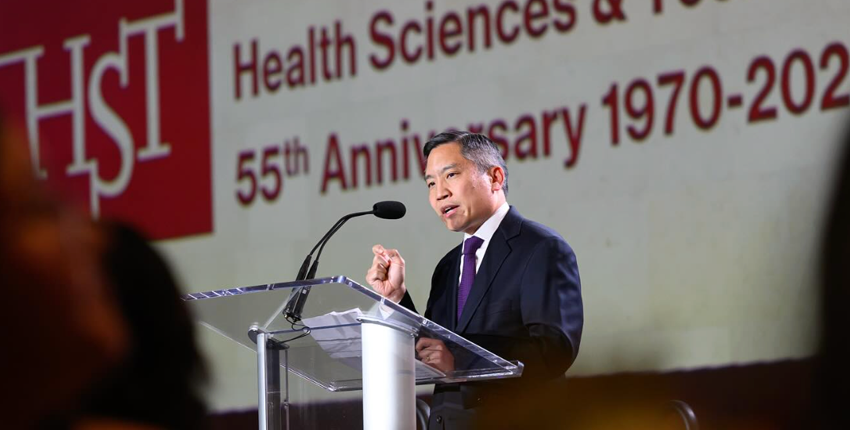 Bernard Chang speaks into a microphone in front of a projection with the HST anniversary dates