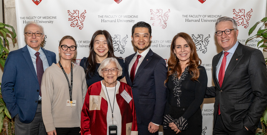 A group of seven adults stands in front of a backdrop displaying the Harvard University Faculty of Medicine logo. They are smiling and posing for a group photo. The group includes an older woman in a red and beige jacket standing in front, with three men and three women standing behind her. Everyone is dressed in professional or semiformal attire, and there are potted plants on either side of the group.