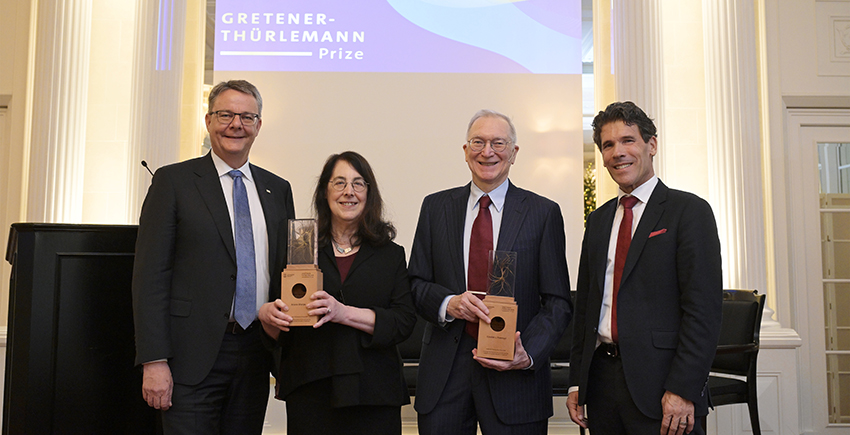 Sharpe and Freeman hold their awards, flanked by Shaepman and Dall'O. They stand in front of a screen with the prize name on it.