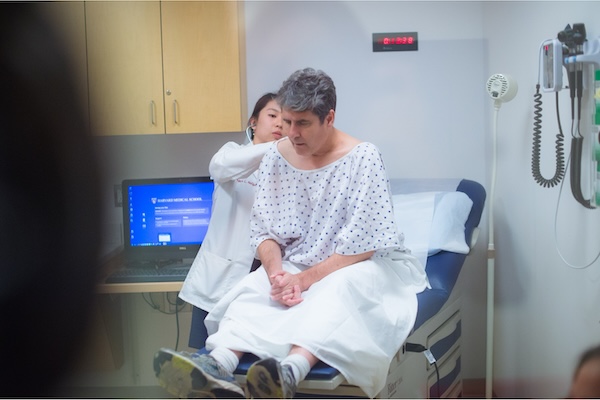 A student stands behind a patient performing a physical exam