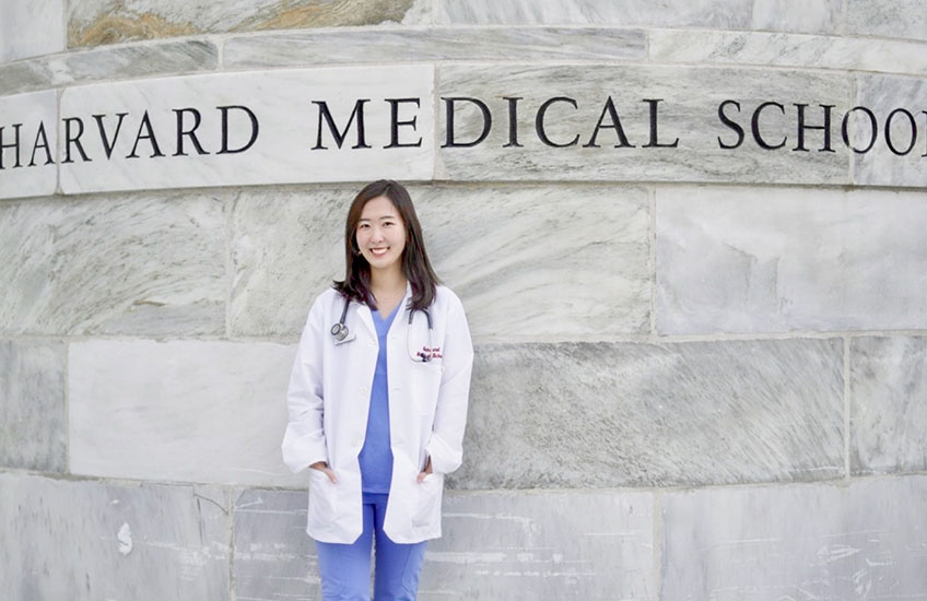 Sarah Lee in scrubs and a white coat standing in front of a marble wall that reads Harvard Medical School.