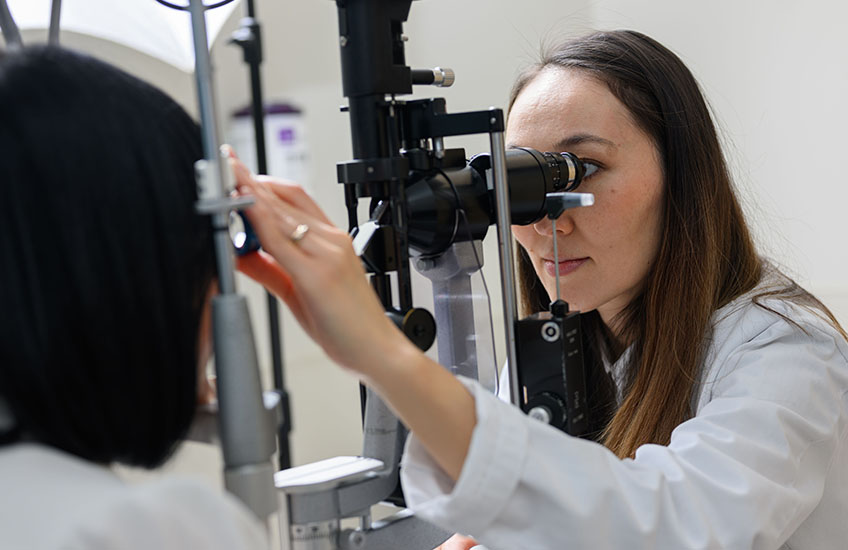 A woman looks through an ophthalmoscope at a patient’s eye while adjusting focus.