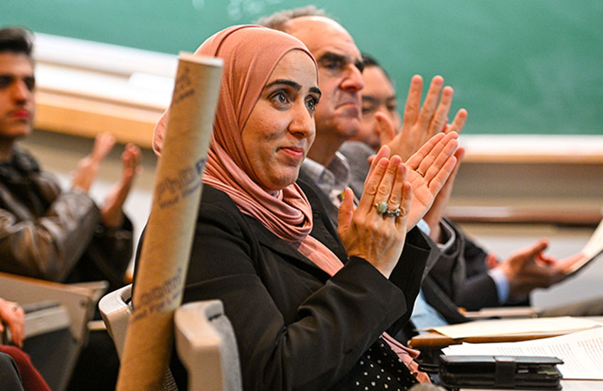 A woman in a pink hijab and black blazer applauds along with other audience members.