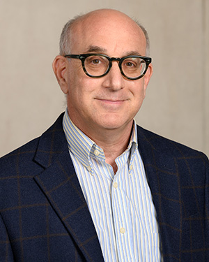 Head shot of a grey-haired man in blue striped oxford and blue suit jacket with glasses