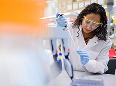A woman in a white coat, exam gloves, and safety goggles pipettes a solution.