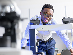 A man in a button shirt and glasses leans over a microscope, smiling