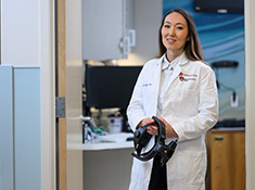 A woman in a white coat stands holding an imaging headset