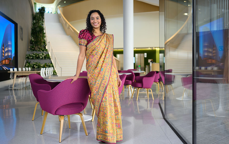 Portrait photo of Anusha Nathan standing alone in an atrium. The gold and fuchsia colors in her sari echo those in the decor of the space.