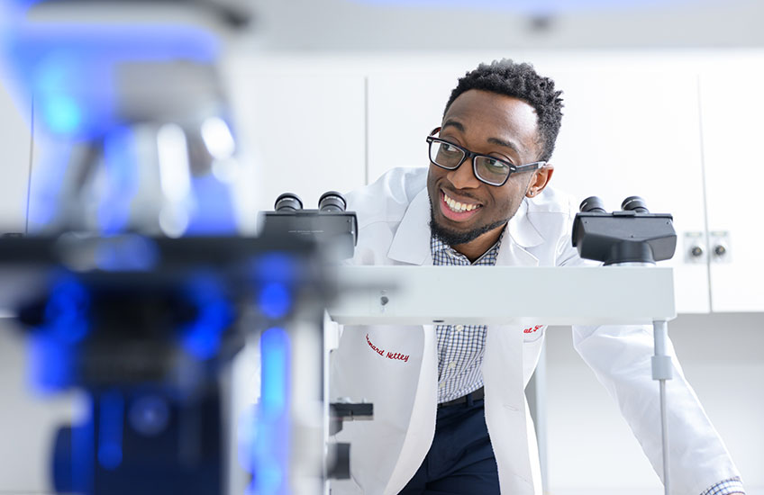 A young man in a white lab coat leans over a pair of microscopes, smiling while looking off to one side.