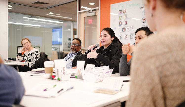 Students and community members sit in groups in a learning studio in discussion.