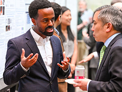 Two men wearing suits engage in conversation in front of a poster.
