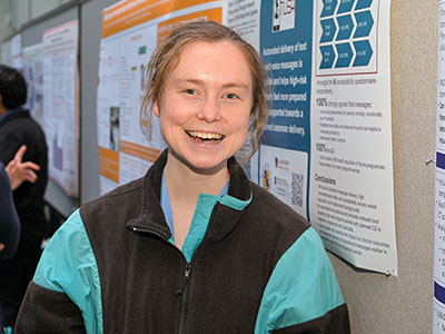 A young woman in a fleece jacket stands in front of a poster, smiling.