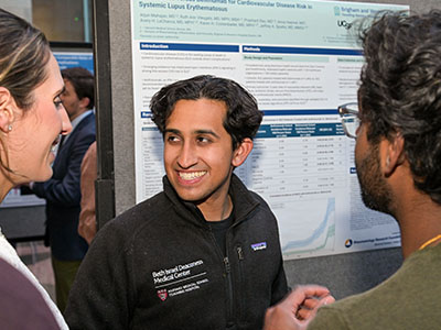 A young man in a fleece jacket smiles at two people. The group is standing in front of a poster.