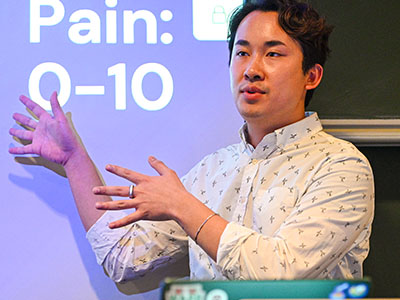 A young man gestures at a large screen while speaking. The screen says “Pain: 0-10.