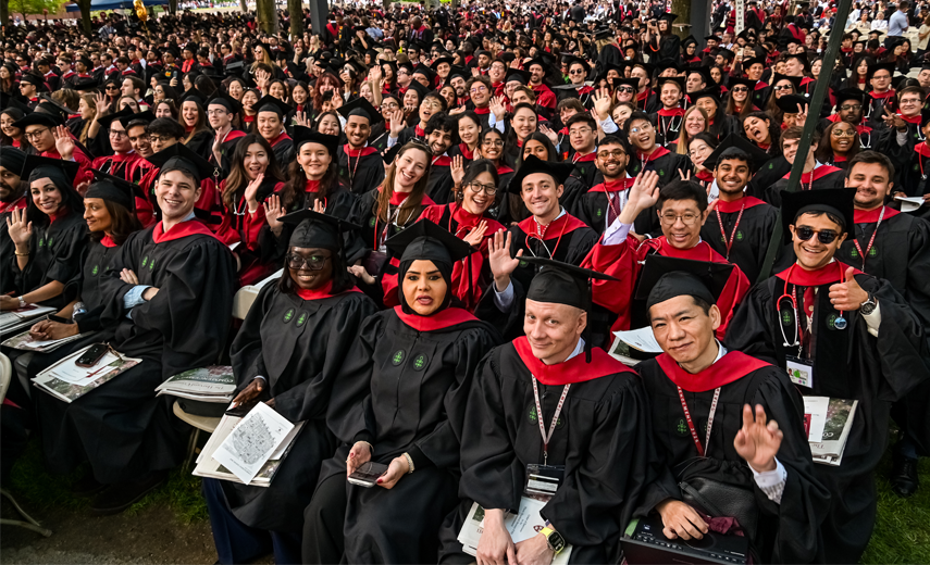 Wide angle photo of a large group of grads in black and red regalia and green crows fee waving and smiling at the camera.