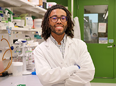 A man in a white coat leans on a lab bench with arms crossed and smiling