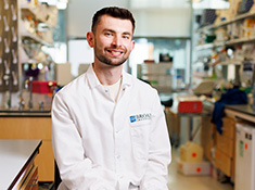 A man in a white lab coat with shelves in a lab in background