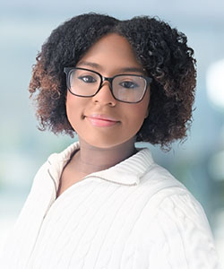 A teenage woman with brown curly hair, wearing glasses and a white sweater, smiles at the camera.