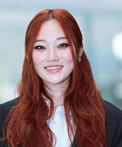 A young woman in a blue suit, with chest-length burgundy-colored hair, stands in a glass-paneled hallway and smiles.