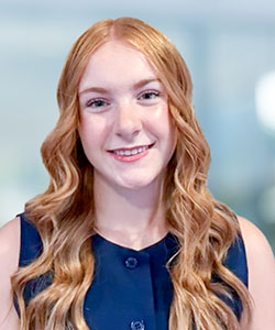 A teenage woman with long, wavy, red hair, wearing a blue sleeveless top, smiles while standing in front of white bricks.