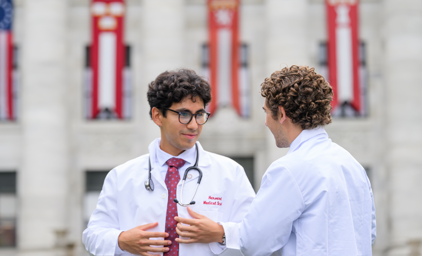 Two new students in white coats converse in front of Gordon Hall, a marble building bedecked with banners.