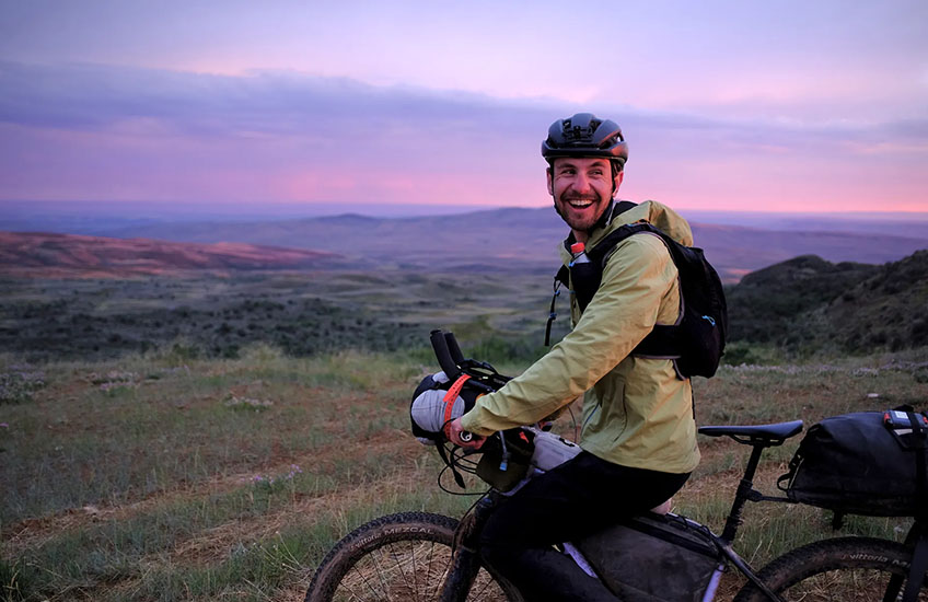 George Usmanov looks back over his shoulder, smiling, while stopped on his bicycle against a grassy landscape at sunrise or sunset.