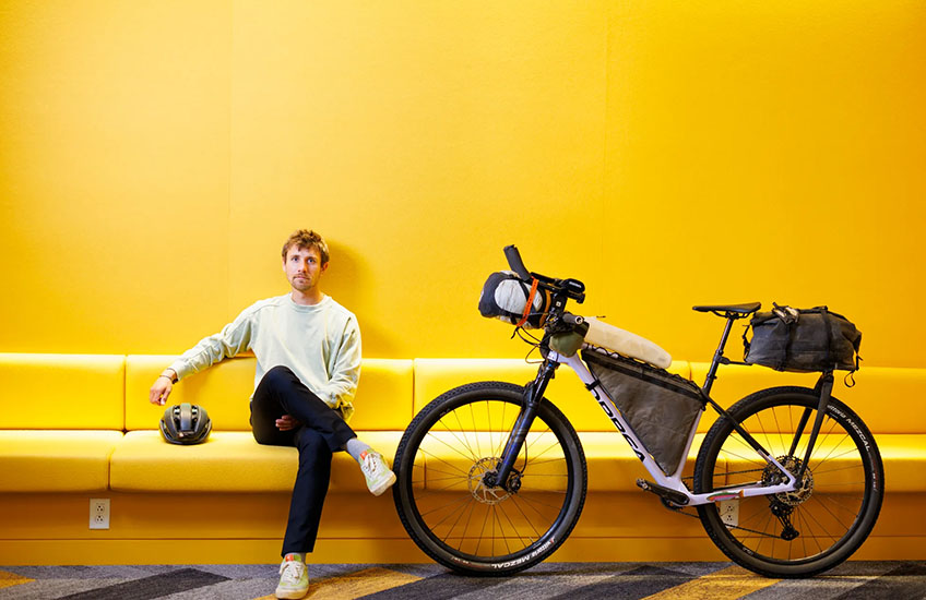 George Usmanov sits on a long yellow cushioned bench next to a bicycle. The wall behind him is also yellow.