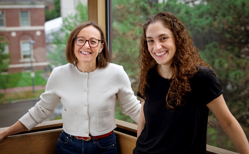 Portrait photo of two women standing in an office corner with large windows overlooking the Harvard campus.