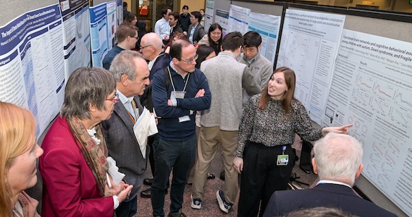 A med student is presenting her research poster to five faculty members gathered around. In the background, other students are also presenting to other attendants.