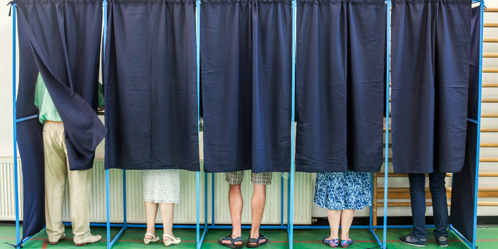 a group of people standing in single voting booths with the curtains down and just their legs and shoes showing.