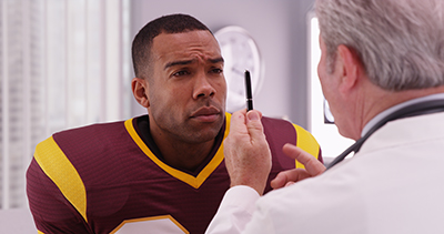 the back of a dr facing a football player squinting looking at the apparatus that the doctor has in his hands. 