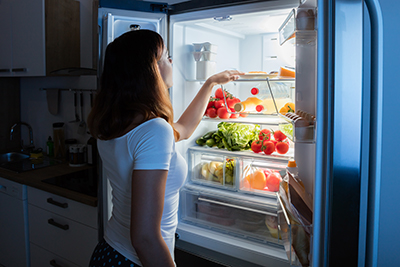 a woman standing in front of the refridgerater in a dim setting with the lights bright from the fridge looking at her food choices. 
