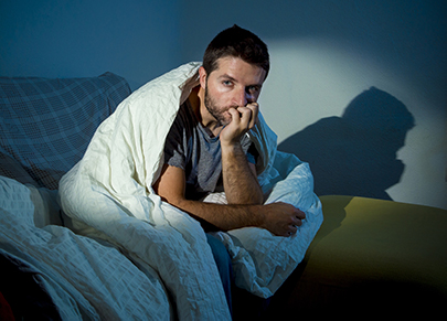 a man sitting on the side of his bed hunched over with his chin resting on his fist and a blanket covering him with a scared look on his face. 